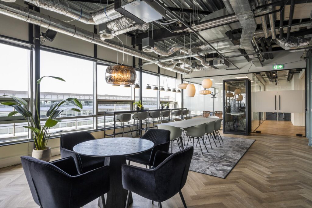 Modern office lounge at Bos en Lommerplein with large windows, stylish seating, and industrial ceiling design.